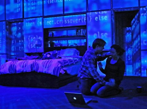 Two figures kneel over a laptop as code is projected across the set